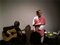 Jamila Ismail reading poetry, seated guitarist to her right
