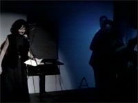 Sheri-D Wilson in front of microphone with guitarist in shadow to her left